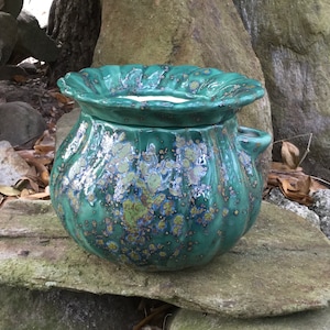 May include: A handcrafted, teal ceramic pot with a ruffled rim and two small handles. The pot has a speckled, iridescent glaze with hints of blue, green, and gold. The pot is sitting on a stone surface.