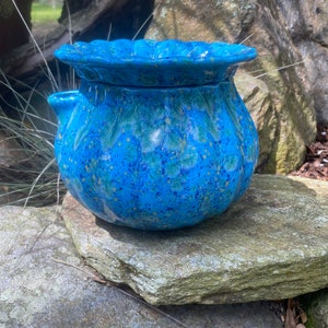 May include: A vibrant blue ceramic pot with a ruffled rim and a textured, speckled surface. The pot has a rounded body and a small spout. It sits on a natural stone surface, suggesting outdoor use. The color palette includes shades of blue and hints of yellow.