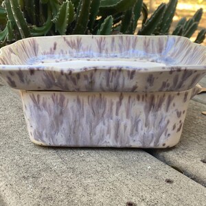 May include: A rectangular ceramic planter with a matching tray. The planter and tray are a light beige colour with a purple, streaked pattern. The planter has a slightly flared rim and is sitting on a concrete surface.