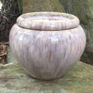 May include: A large, round ceramic jar with a lid. The jar is a mix of beige, brown, and purple hues, with a glossy finish. The lid sits on top, creating a decorative and functional piece. The jar is sitting on a rock.