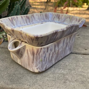 May include: A two-piece ceramic dish set with a rectangular shape. The set features a marbled pattern in shades of beige, purple, and white. The top dish has a wavy rim and a white interior, while the bottom piece has a handle.