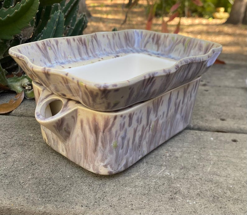 May include: A two-piece ceramic dish set with a rectangular shape. The set features a marbled pattern in shades of beige, purple, and white. The top dish has a wavy rim and a white interior, while the bottom piece has a handle.