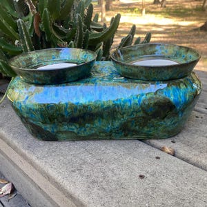 May include: A ceramic pet bowl set with two round bowls and a rectangular base. The bowls and base have a glossy, marbled finish in shades of blue, green, and brown. The bowls are designed to hold water or food for pets.