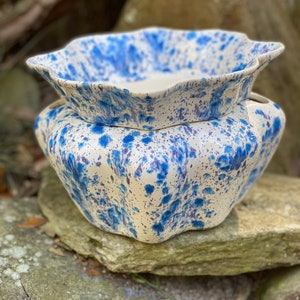 May include: Two ceramic bowls, one stacked on the other, with a cream-colored base and blue splatter design. The top bowl has a scalloped edge. The bowls are sitting on a stone surface.