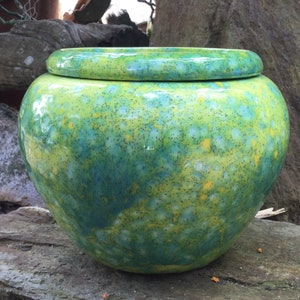 May include: A round, ceramic jar with a lid, featuring a speckled glaze in shades of green, yellow, and blue. The jar has a glossy finish and sits on a stone surface. The jar is a decorative item.