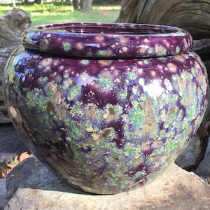 May include: A round, glazed ceramic pot with a lid. The pot is a deep purple color with speckled green, blue, and gold accents. The pot sits on a stone surface and has a glossy finish. The pot is a decorative item.