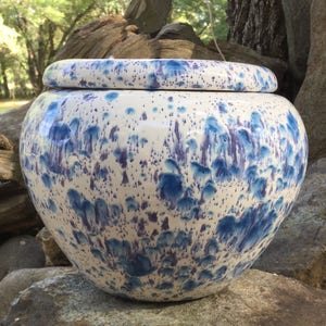 May include: A ceramic jar with a lid, featuring a speckled design in shades of blue and purple against a white background. The jar has a rounded shape and a matching lid. The piece is likely handmade and suitable for decorative or functional use.