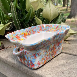 May include: A rectangular ceramic planter with a white interior and a colorful exterior. The exterior features a speckled pattern of blue, orange, and red. The planter has two small handles and is suitable for small plants or succulents.