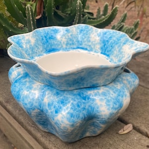 May include: Two-piece ceramic serving dish set. The set features a wavy, scalloped edge design. The exterior is a speckled blue and white pattern, while the interior is white. The dishes are stacked on a wooden surface.