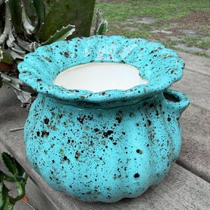 May include: A turquoise ceramic planter with a ruffled, scalloped rim and a white interior. The exterior features a speckled pattern of black and dark brown spots across its rounded, ribbed body, which includes a small side opening for drainage.