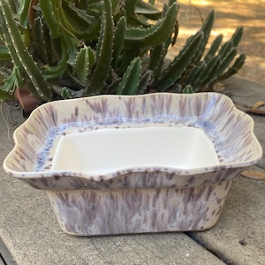 May include: A ceramic serving dish with a rectangular white insert. The outer dish has a wavy rim and a speckled purple and cream glaze. The dish is set against a backdrop of green plants and a weathered wooden surface.