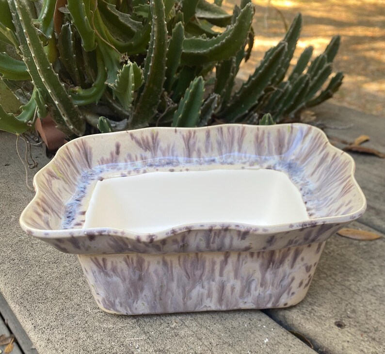 May include: A ceramic serving dish with a rectangular white insert. The outer dish has a wavy rim and a speckled purple and cream glaze. The dish is set against a backdrop of green plants and a weathered wooden surface.