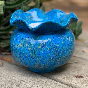 May include: A round ceramic planter with a ruffled, wavy rim. The exterior features a vibrant blue glaze with a speckled, mottled pattern in shades of lighter blue, green, and yellow. The interior of the bowl is white and smooth.