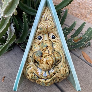 May include: A ceramic wall hanging featuring a whimsical face with a prominent nose and textured details. The face is primarily tan with green and brown accents, framed by a light blue triangular border. A metal wire is attached for hanging.