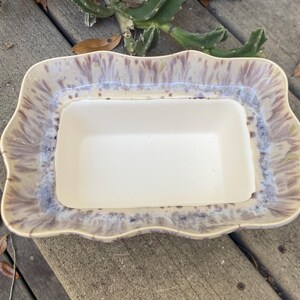 May include: A rectangular ceramic serving dish with a scalloped edge. The dish has a white interior and a rim with a blend of beige, purple, and blue hues. The dish is likely used for serving food.