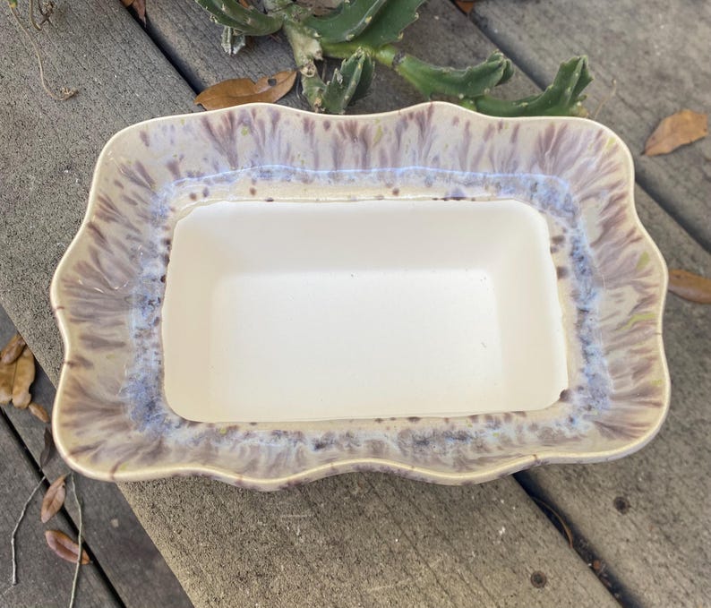 May include: A rectangular ceramic serving dish with a scalloped edge. The dish has a white interior and a rim with a blend of beige, purple, and blue hues. The dish is likely used for serving food.