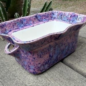 May include: A rectangular ceramic planter with a wavy rim. The planter has a speckled pink and blue glaze on the exterior and a white interior. It features two small handles on the sides, and is designed for planting flowers or herbs.