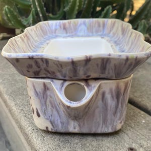 May include: A two-piece ceramic wax warmer with a speckled purple and white glaze. The top dish has an irregular shape and a white interior. The base has a spout and a circular opening. Designed for melting wax melts, it measures approximately 15 cm in height.