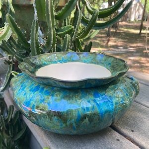 May include: A two-piece ceramic planter set with a glossy, marbled glaze in shades of blue and green. The larger base has a rounded shape with handles, and the smaller top piece has a scalloped edge and a white interior. The set is designed for plants.