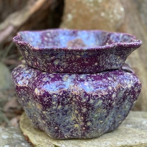 May include: A two-piece ceramic planter set with a deep purple glaze and speckled, mottled texture. The top piece is a shallow dish with a wavy rim, and the bottom is a bulbous, star-shaped pot. The set is designed for indoor or outdoor use.