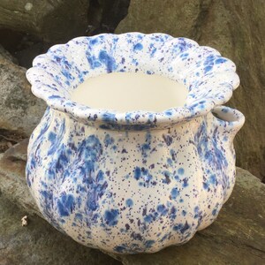 May include: A ceramic planter with a scalloped rim and a speckled blue and white design. The planter has a rounded shape with a small handle on the side. The interior is a solid cream color.