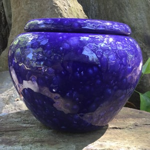 May include: A round, deep blue ceramic jar with a matching lid. The jar has a glossy finish with a textured, speckled pattern. The jar is sitting on a stone surface.