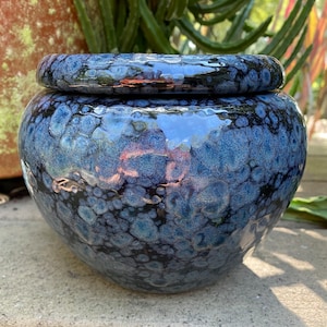 May include: A round, ceramic jar with a lid, featuring a speckled, glossy blue glaze. The jar's surface has a textured appearance with dark blue and black spots. The lid sits atop the jar, reflecting light. This decorative jar is suitable for storage or display.