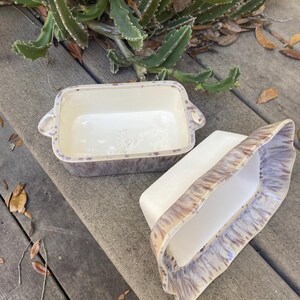 May include: Two rectangular ceramic baking dishes. One is upright, the other is on its side. Both have a white interior and a speckled, purple and brown exterior. The dishes have handles on the sides.