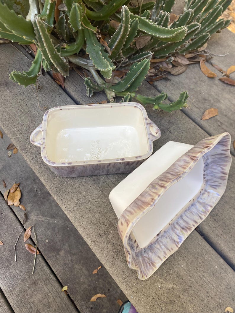 May include: Two rectangular ceramic baking dishes. One is upright, the other is on its side. Both have a white interior and a speckled, purple and brown exterior. The dishes have handles on the sides.