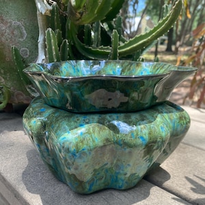 May include: Two-piece ceramic pottery set with a glossy, marbled finish in shades of green and blue. The top piece is a shallow, scalloped dish with a dark rim, resting on a larger, textured base. The set is designed for decorative or functional use.