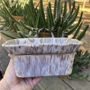 May include: Two ceramic rectangular baking dishes with a lavender and cream marbled glaze. The dishes have a slightly scalloped edge and are stacked. The background features green plants and a wooden surface.