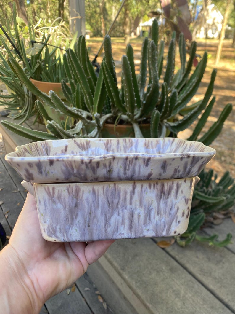 May include: Two ceramic rectangular baking dishes with a lavender and cream marbled glaze. The dishes have a slightly scalloped edge and are stacked. The background features green plants and a wooden surface.