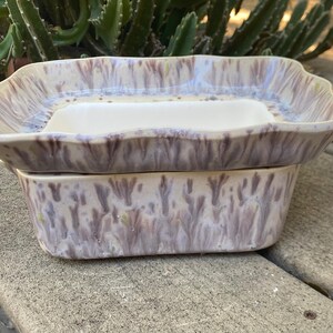May include: A two-piece ceramic soap dish set. The top piece is a rectangular dish with a raised rim and a white interior. The bottom piece is a matching rectangular base. The set has a lavender and cream glaze.