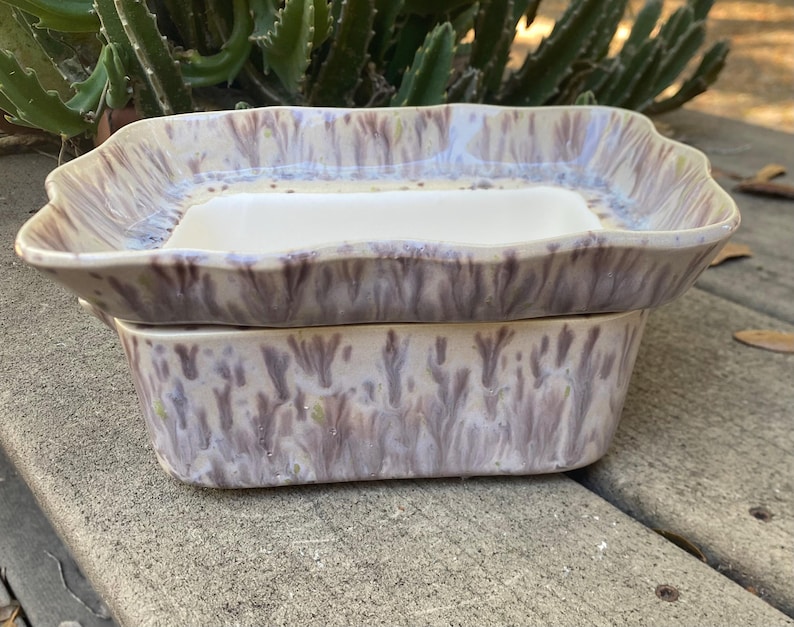 May include: A two-piece ceramic soap dish set. The top piece is a rectangular dish with a raised rim and a white interior. The bottom piece is a matching rectangular base. The set has a lavender and cream glaze.