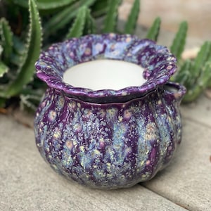 May include: A ceramic planter with a textured, speckled glaze in shades of purple, blue, and yellow. The planter has a ruffled rim and a round body. The interior is white. The planter is suitable for small plants.
