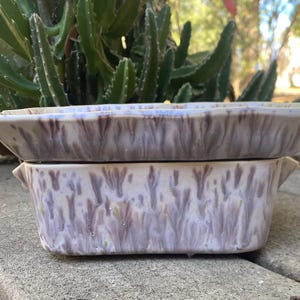 May include: Two ceramic rectangular planters stacked. The planters have a speckled glaze in lavender, brown, and cream. The top planter has a different shape. The planters are on a concrete surface. The planters are suitable for small plants or succulents.
