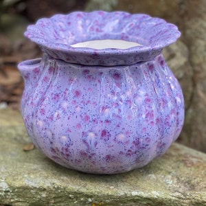 May include: A speckled lavender ceramic wax warmer with a scalloped rim and two small handles. The warmer has a bulbous shape and a white interior. The exterior is decorated with dark pink spots.