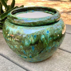 May include: A round, ceramic planter with a glossy, mottled glaze in shades of green and blue. The planter has a wide, circular rim and a matching lid. The surface reflects light, highlighting the color variations. Ideal for indoor or outdoor use.