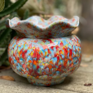 May include: A ceramic container with a lid, featuring a vibrant speckled design in shades of blue, red, orange, and yellow. The lid has a ruffled edge. The container is round with a pedestal base.