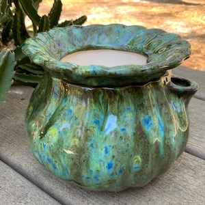 May include: A handcrafted ceramic planter with a glossy, textured finish. The planter features a ruffled rim and a small spout. The exterior is a blend of green and blue hues, with a white interior. The planter is designed for indoor or outdoor use.
