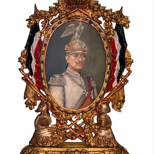 Puede incluir: Un retrato antiguo en un marco dorado ornamentado. El retrato ovalado representa a un hombre con uniforme militar, coronado con un casco y un águila. El marco presenta banderas en negro, blanco y rojo, con elementos decorativos.