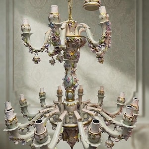 May include: A multi-tiered porcelain chandelier featuring an off-white finish with gold accents and intricate floral embellishments. The fixture has numerous light sockets arranged on curved arms, decorated with sculpted roses and pastel-colored petals.