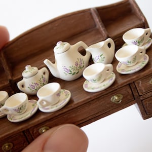 May include: A miniature tea set featuring a teapot, creamer, sugar bowl, and six cups and saucers. The set is decorated with a floral pattern in shades of purple and green. The set is displayed on a wooden table with drawers.