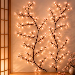 以下が含まれることがあります： 壁掛け式の桜の枝を模した装飾用ライトストリング2本。濃い茶色の枝には、半透明のピンク色の花びら型LEDライトが多数付いており、壁に向かって温かみのある柔らかな光を放っています。