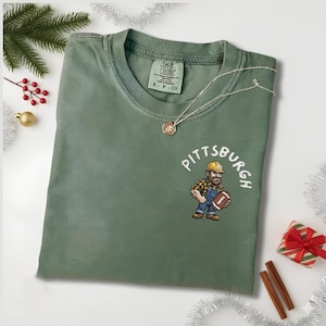 May include: An olive green Comfort Colors t-shirt with a crew neck and short sleeves. The shirt features an embroidered design of a construction worker holding a football, with the word "Pittsburgh" above. A silver necklace with a pendant is also visible.