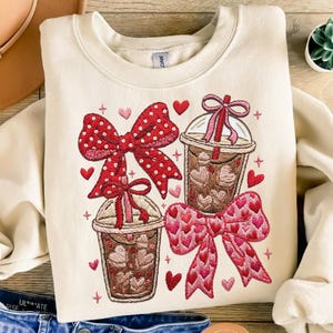 May include: Cream-coloured sweatshirt with embroidered design featuring two iced coffee cups and two bows. One bow is red with white polka dots, the other is pink with heart patterns. Small red hearts and pink crosses are scattered around the design.