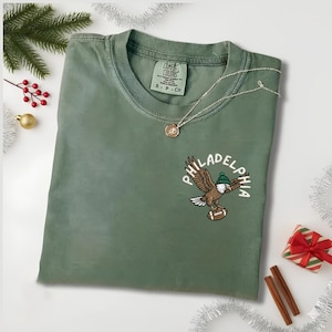 May include: An olive green t-shirt with a graphic of an eagle wearing a green hat and holding a football, with the word "PHILADELPHIA" in a curved design. A silver necklace with a pendant is on the shirt.