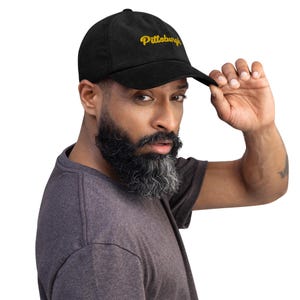 Può includere: Un berretto da baseball in velluto a coste nero con un logo "Pittsburgh" ricamato in oro. Il berretto è indossato da una persona con la barba.