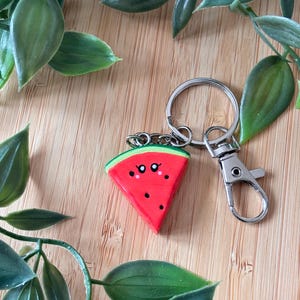 Op de afbeelding: Een sleutelhanger met een watermeloen schijfje en een zilveren sluiting. De sleutelhanger heeft een rode watermeloen schijf met zwarte zaden, een groene schil en twee kleine zwarte ogen. De sleutelhanger ligt op een houten oppervlak met groene bladeren.