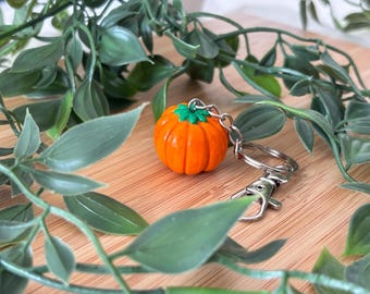 Può includere: Un portachiavi a forma di zucca arancione con uno stelo verde. Il portachiavi ha un moschettone e un anello argentati. La zucca è circondata da foglie verdi e posta su legno.